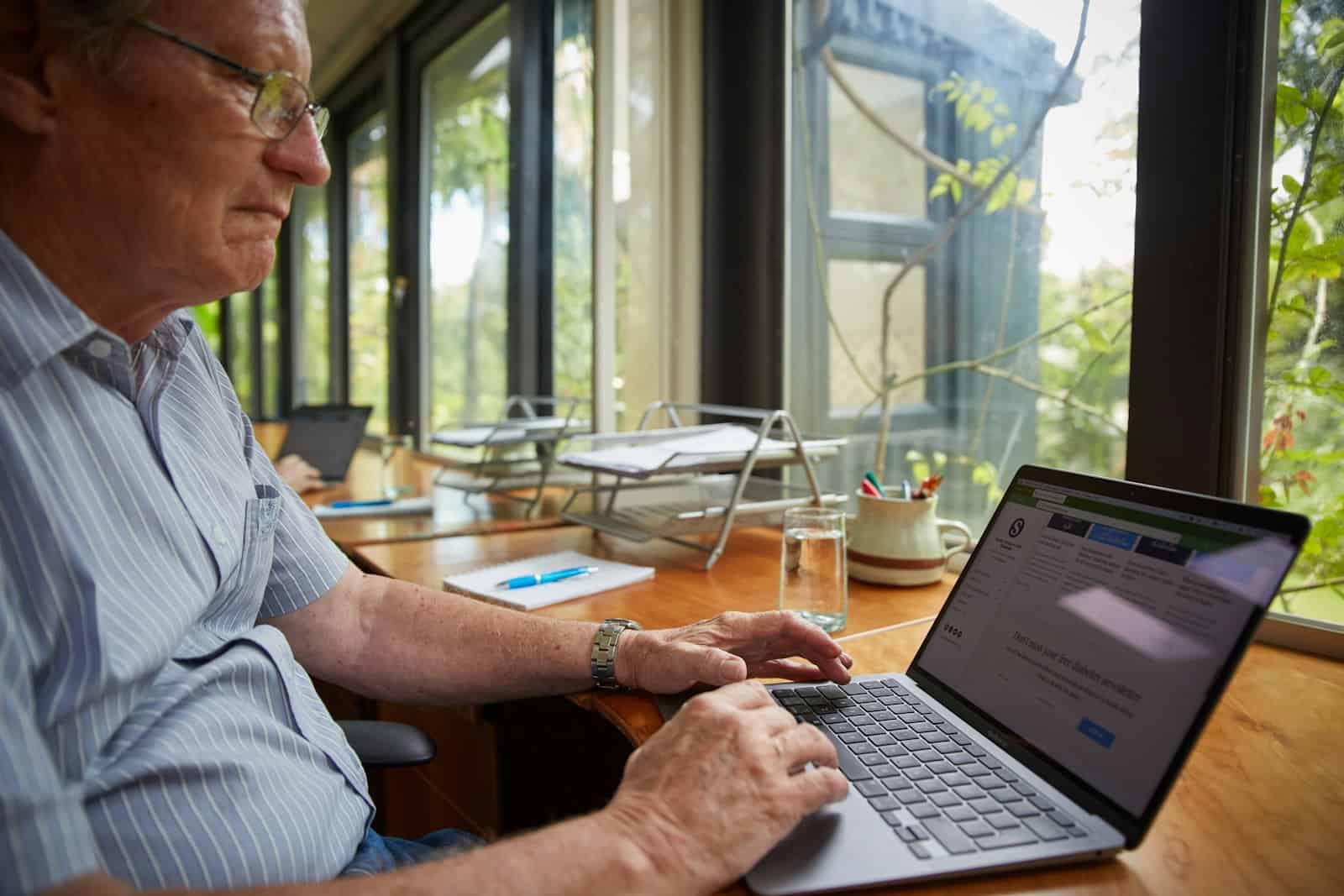 An older man using a laptop at a table in his home office.