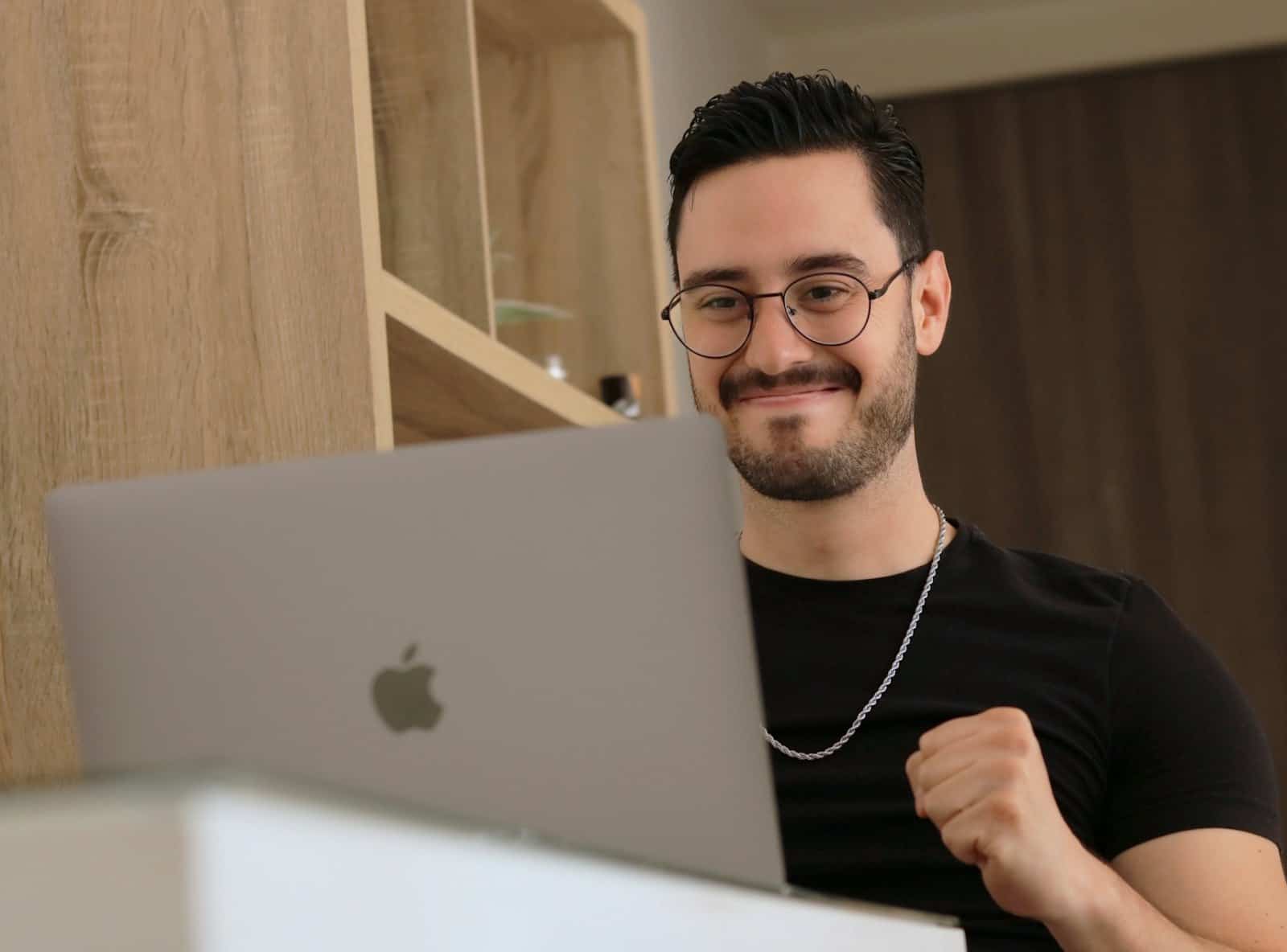A smiling man in glasses working on a laptop at home.