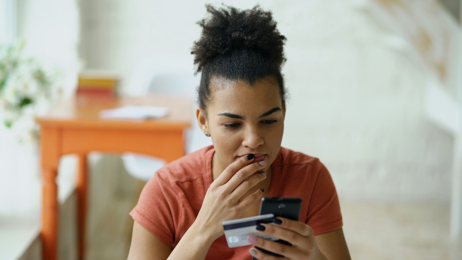 A smiling young woman at home looks at a banking app on her phone while holding a credit card.