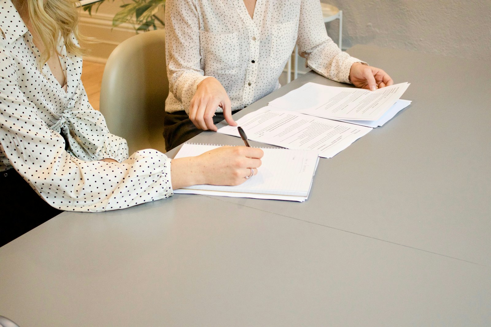 A woman signs a document at a desk while another woman reaches for the papers during an appointment.