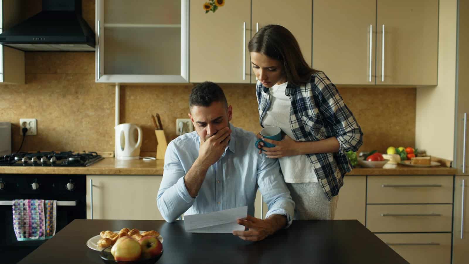 A young man reads a stack of bills at the kitchen table while his partner hugs him.