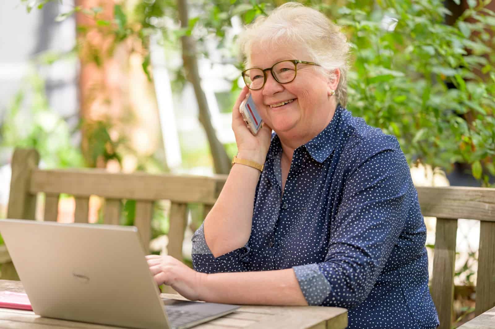 An older woman in glasses talks on her phone while working on a laptop at a table.