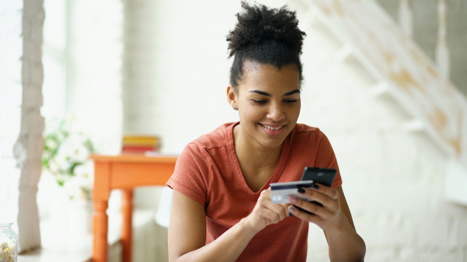 A young woman holds a bank card and her phone while checking an account at home.