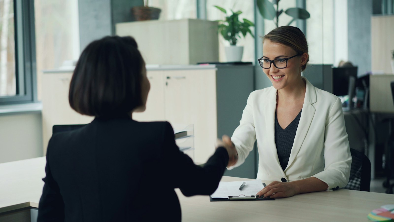 Two women shake hands across an office desk during a hiring meeting.