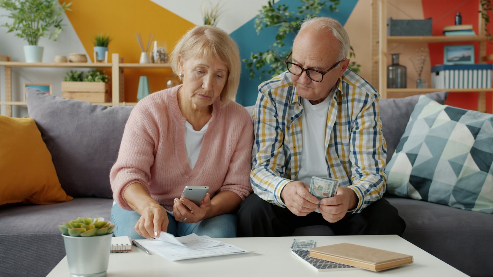 An older couple sits at home going over bills and financial paperwork with a phone.