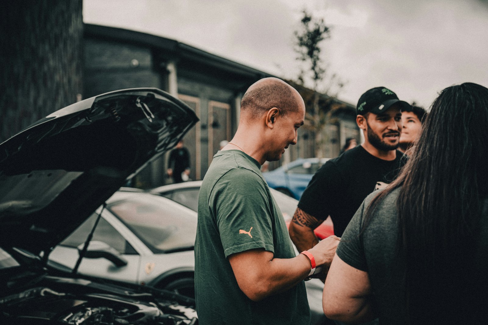 A man stands beside a car with its hood open, talking with others about an unexpected repair.