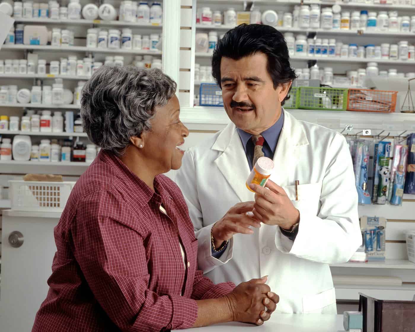 A pharmacist holds up a pill bottle and explains a prescription to an older woman at the pharmacy counter.