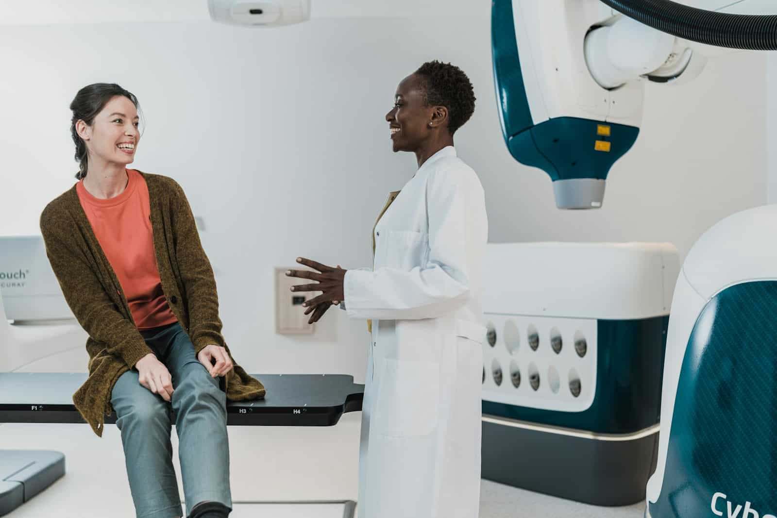 A doctor in a white coat talks with a patient in an exam room.