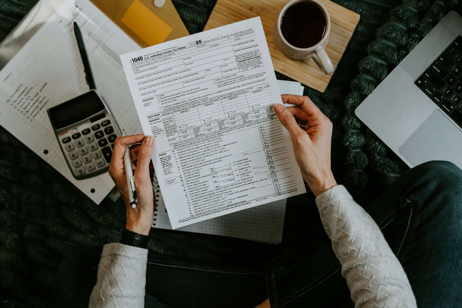 Hands holding tax forms next to a calculator, pen and laptop on a home desk.