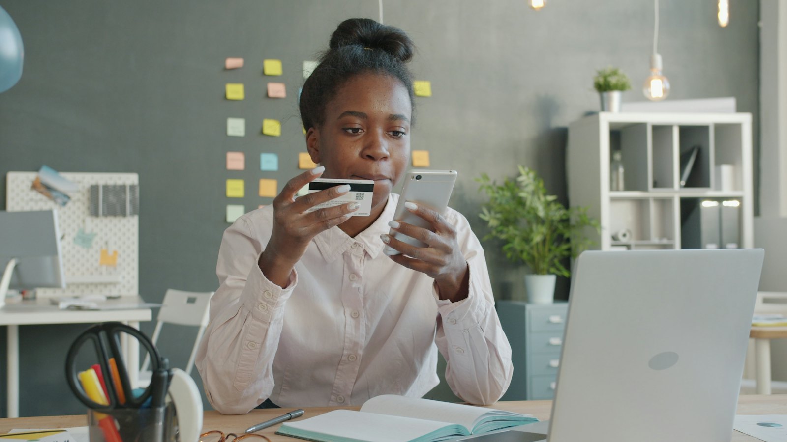 A smiling woman at a desk holds a bank card while using her smartphone.