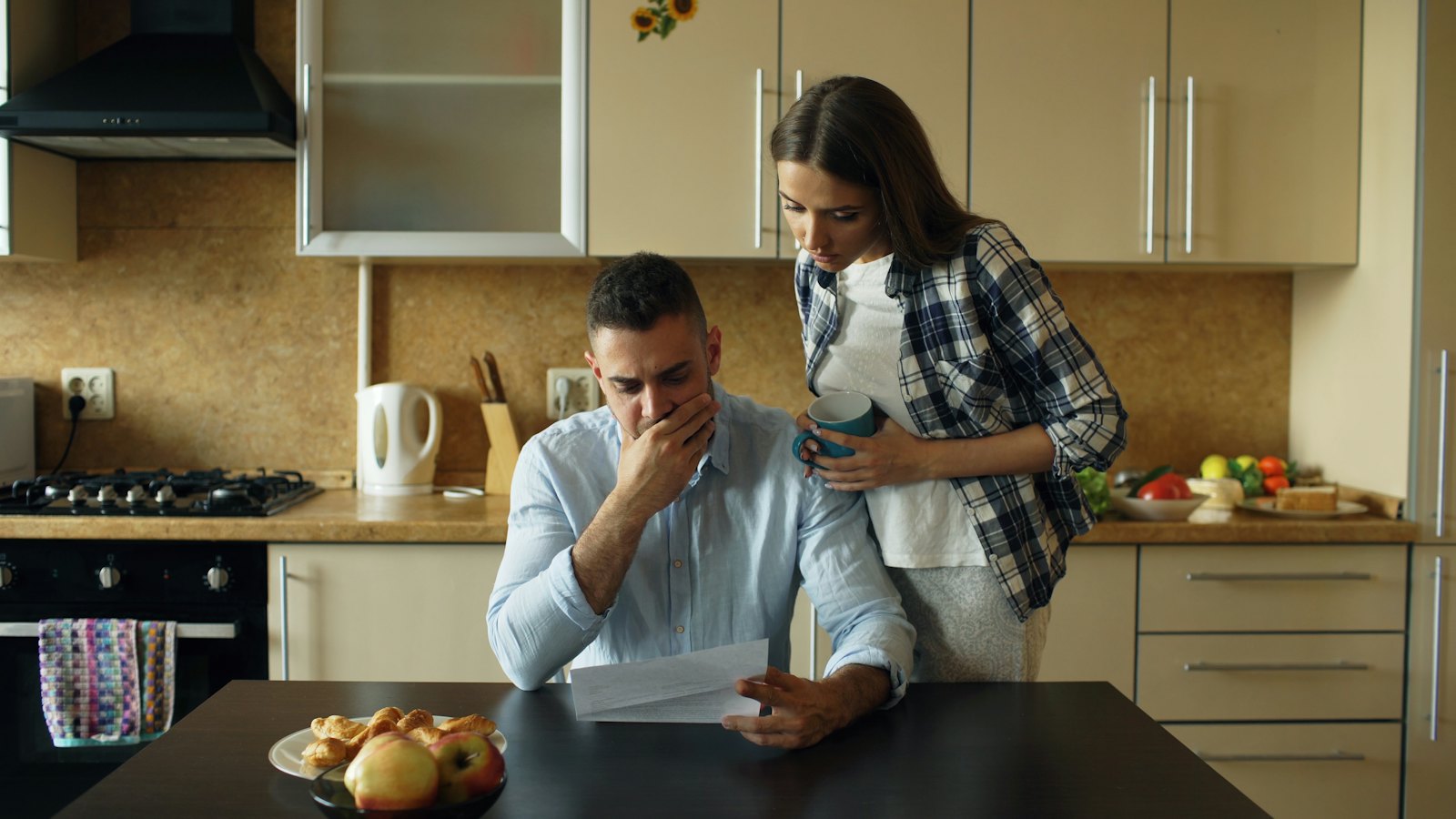 A worried young man sits at the kitchen table with a stack of bills while his wife comforts him.