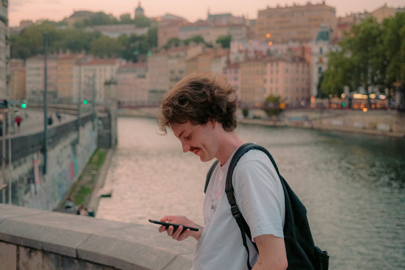 A young man sits on a ledge outdoors, looking down at his phone.