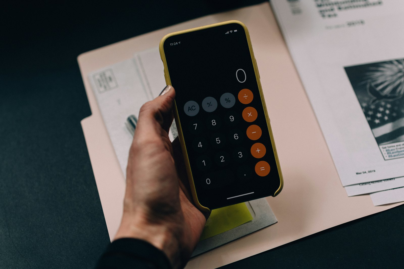 A woman holds a smartphone with a calculator app open over a paper folder and a cup of coffee